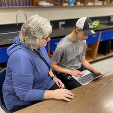 Female Education Assistant sits with male secondary student, helping with an assignment
